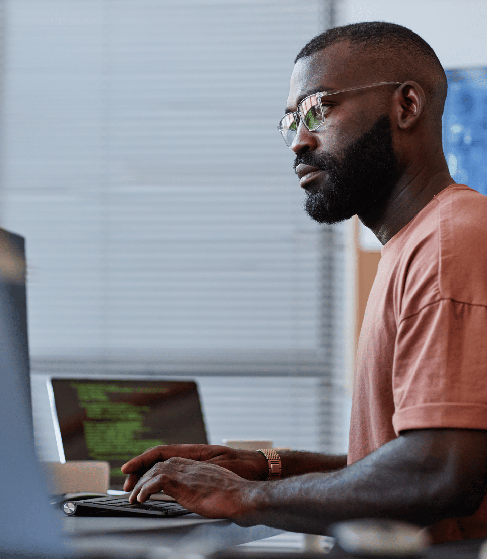 Young IT Professional with Glasses Working on Laptop with Screens in the Back